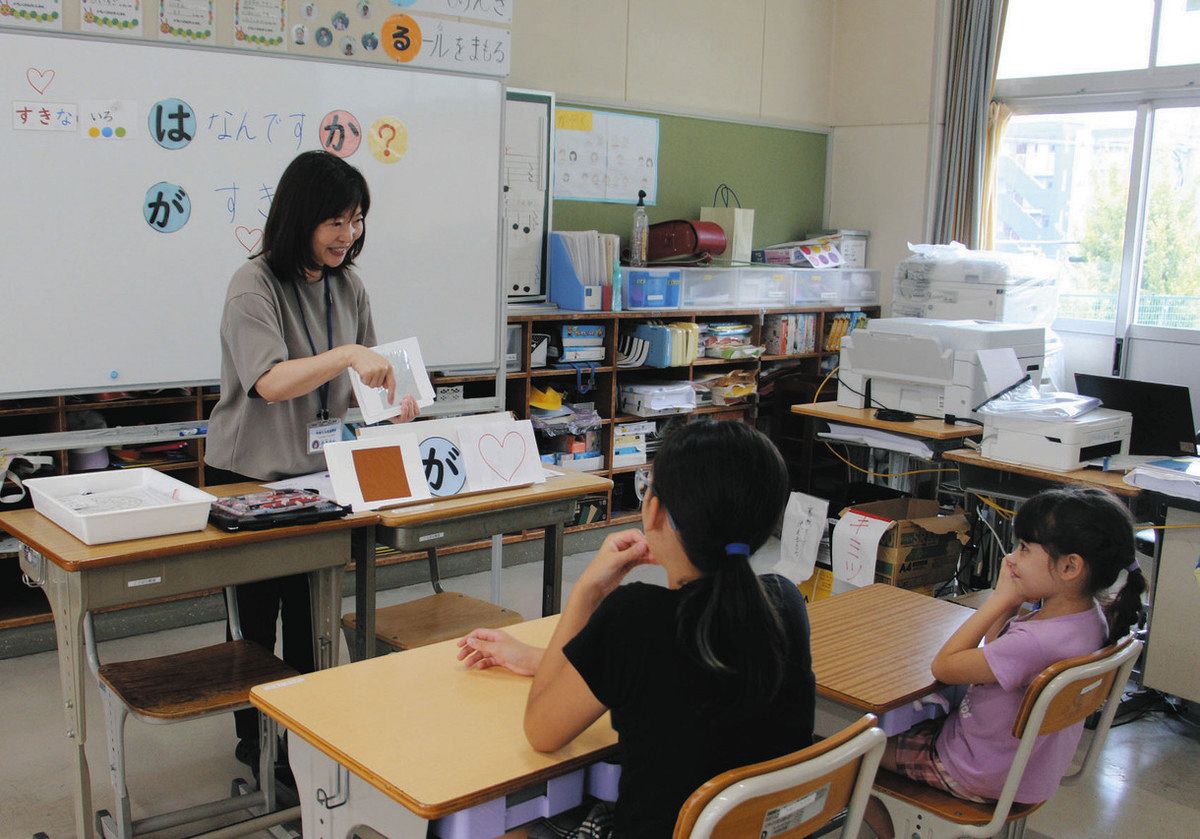 指導員（左）が多彩な色の紙を示し「ちゃいろ」「ぎんいろ」などと答える児童たち＝愛知県豊田市の西保見小で
